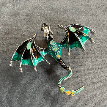 Das Produktbild zeigt eine Brosche in Form eines Drachen, mit ausgebreiteten Flügeln. Auf dem Rücken und dem Schwanz sind jeweils drei helle runde Edelsteine, auf den Flügeln jeweils ein türkiser ovaler Edelstein. Der Drache ist türkis, schwarz und silber.